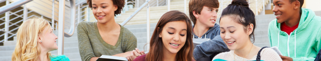 Six high school students sitting outside on steps smiling, talking, and looking at books and laptops.