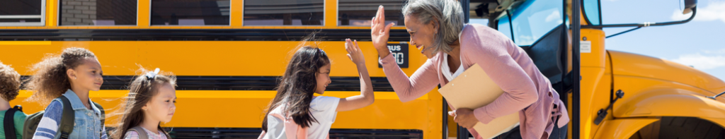An African American teacher high-fives a young female student in line for the school bus as other students wait in line for their turn.