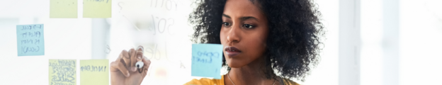 An African American woman brainstorms plans by writing with a dry-erase marker on a glass wall that also includes sticky notes.