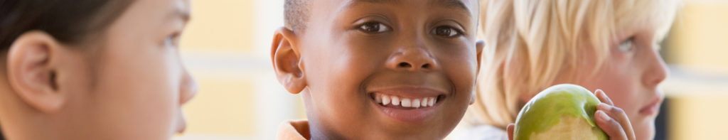 A Black male elementary student smiles while holding a green apple.