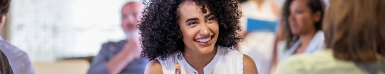 Young woman with dark curly hair smiles as she talks with another woman who is facing her. Other people talk with other in the background.