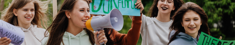 Diverse group of teens rally for stopping climate change; female student at the front of the group speaks into a megaphone while others hold signs.