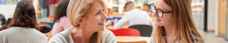 A white female teacher listens to a white female high school student, both are smiling and engaged.