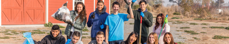 Diverse group of teenagers holding filled trash bags, yard tools, and other cleaning materials smiling at their successful community pickup.