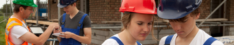 Two teens wearing coveralls and hardhats discuss plans while the construction teacher talks with another students; unfinished construction frame in the background.