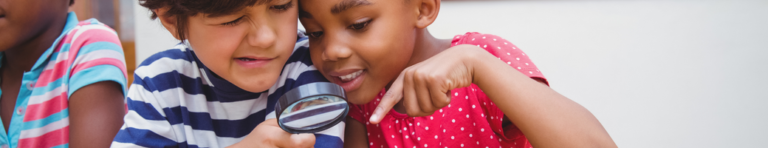 White male and Black female elementary students look through a magnifying glass to examine an object not shown in the image.