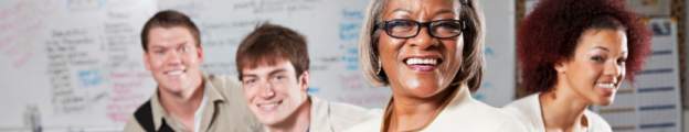 Black female teacher wearing glasses smiles at the camera with three smiling high school students behind her.