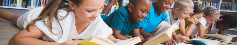 Group of seven diverse elementary student lying on their stomachs on the library floor and smiling while they each read a different book.