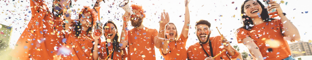 Group of high school students all dressed in orange tee-shirts and orange face paint cheering their team while multi-colored confetti falls around them.