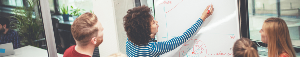 Young Black woman with natural hair marks a point on a grid chart on a whiteboard with a red dry erase marker as three colleagues observe.