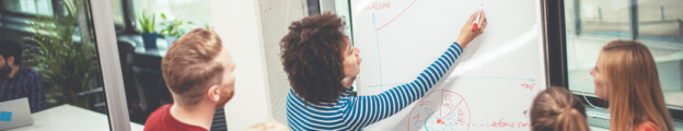 Young Black woman with natural hair marks a point on a grid chart on a whiteboard with a red dry erase marker as three colleagues observe.