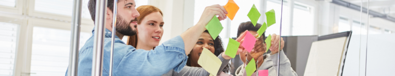 Group of four diverse professionals looking at and pointing to sticky notes on a glass wall.