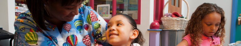 A Black female nurse wearing scrubs with colorful hot air balloons comforts a young girl in a hospital children's area.