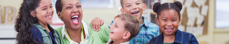 Black female teacher with short haircut and green cardigan laughs with a diverse group of elementary students.