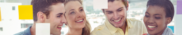 Four young adults smile as they look at a glass wall of yellow sticky notes.