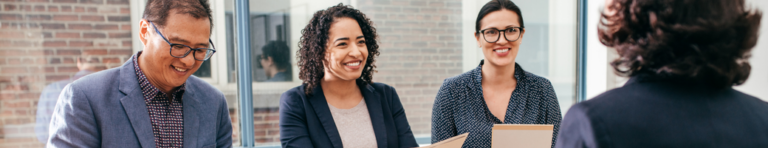 A diverse interview panel of three casual professionals smiles at a job candidate with medium length dark hair who is only seen from the back.