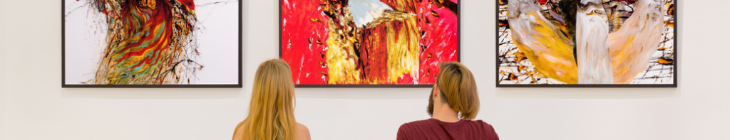 Two people facing away from the camera look at a wall with three large paintings hung on it.