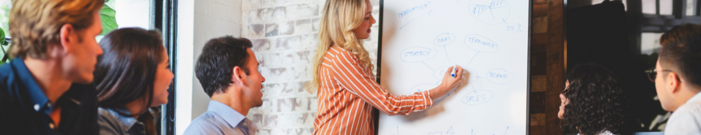 Blonde woman brainstorms ideas on a large whiteboard that are presented from a group of diverse casual professionals sitting around a large meeting table.