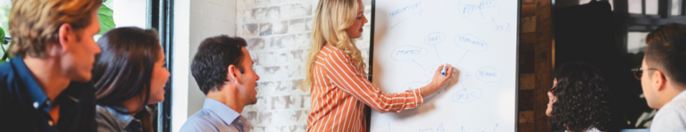 Blonde woman brainstorms ideas on a large whiteboard that are presented from a group of diverse casual professionals sitting around a large meeting table.