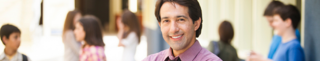 White male school principal smiling with students on campus out of focus in the background.