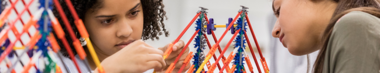 Two middle school girls with dark complexions and features build a bridge with architecture building toys.