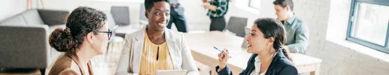 Three diverse professional women sit at a table and discuss ideas.