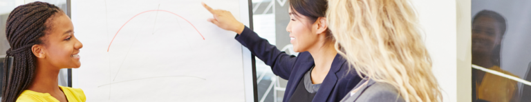 Three diverse professional woman look at a poster paper with one pointing at it.