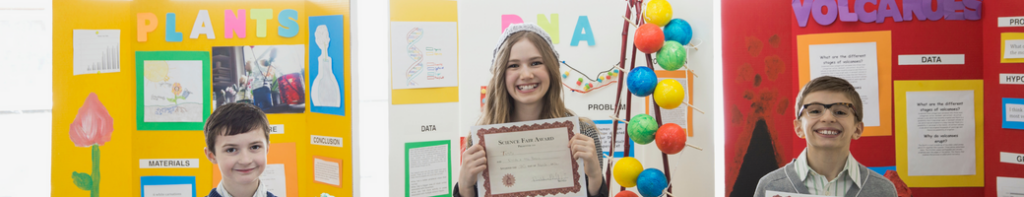 Three smiling, older elementary school students holding certificates and standing in front of their respective science fair poster-board displays.