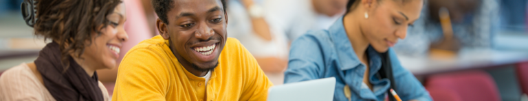 Smiling Black female and male high school students work together at a laptop. while other students work independently in the background.