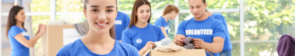High school students in blue t-shirts with the word "volunteer" printed on them sort donated clothes in cardboard boxes. One female student faces the camera smiling.