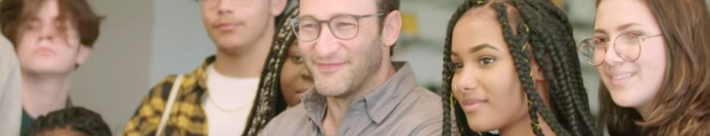A smiling Simon Sinek surrounded within a standing group of diverse students, most prominently an African American teen girl to the right.