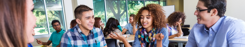 Diverse group of four high school students have a lively conversation while other small groups of students in the background do the same.