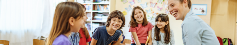 A female teachers and a diverse group of elementary students sit on desktops in a circle and laugh with each other.