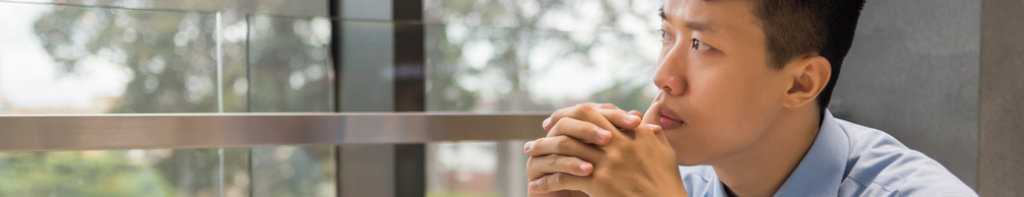 Asian-presenting male adult professional viewed at profile looking out a window reflectively with his clasped hands at his chin and mouth.